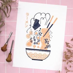 Könnte beinhalten: Ein weißer Druck mit einer schwarzen und orangefarbenen Illustration einer Schüssel Ramen. Der Text "Ramen" ist in japanischen Schriftzeichen auf dem Druck geschrieben.