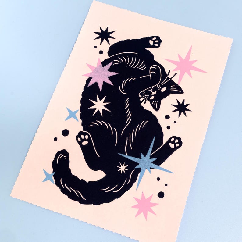 Puede incluir: Una l&aacute;mina de arte rectangular sobre papel rosa claro que presenta la silueta de un gato negro en una pose juguetona. El gato est&aacute; rodeado por varias formas de estrellas en tonos azul oscuro, azul claro y rosa, junto con peque&ntilde;os puntos negros.