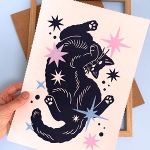 Puede incluir: Una l&aacute;mina de arte rectangular con borde festoneado que muestra un gato azul marino oscuro en una posici&oacute;n juguetona. El gato est&aacute; rodeado de varias formas de estrellas en tonos rosa claro, azul marino oscuro y azul claro sobre un fondo crema.