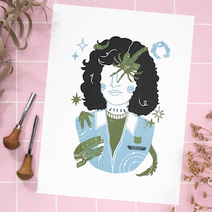 May include: A blue and green print of a person with a creature from the movie Alien on their shoulder. The person has black hair and a green shirt. The creature has a long tail and is holding onto the person's shoulder with its arms.