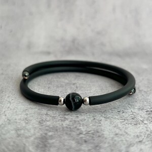 Puede incluir: Una pulsera de estilo envolvente con una banda negra mate lisa, una cuenta central redonda de ágata con bandas blancas y negras, y pequeñas cuentas espaciadoras metálicas de tono plateado a ambos lados de la piedra.