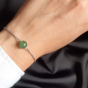 Puede incluir: Una delicada pulsera con una pequeña gema verde redonda. La gema es el punto focal, conectada a una fina cadena plateada. La pulsera se muestra en una muñeca, sobre un fondo oscuro.