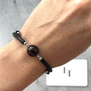 May include: A black bracelet with a large, round, dark red gemstone bead. The bracelet has black rubber or silicone bands and silver-coloured metal accents. The bracelet is worn on a wrist.