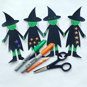 May include: Four paper witch cutouts in black dresses and hats, holding hands. Green and orange Sharpie markers, a pencil, and scissors are arranged near the witches. The witches have green faces and star accents on their dresses. A fun Halloween craft.