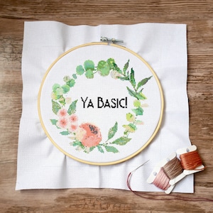 May include: A white embroidery hoop with a floral wreath design and the text "YA BASIC!" in black. The wreath is made of green leaves and pink flowers. The hoop is on a white fabric background.