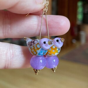 May include: Purple glass bead earrings with a pair of small, colorful glass birds attached to the top of each bead.