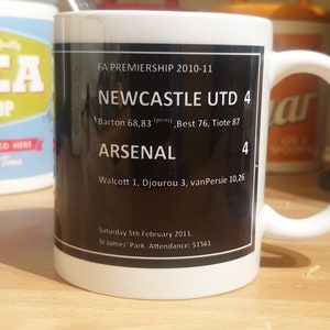 K&ouml;nnte beinhalten: Eine wei&szlig;e Keramiktasse mit einem schwarzen Design, das den Text "FA Premiership 2010-11", "Newcastle Utd 4", "Arsenal 4" und die Ergebnisse des Spiels zeigt. Der Text "Saturday 5th February 2011. St James' Park. Attendance: 51561" ist ebenfalls enthalten.