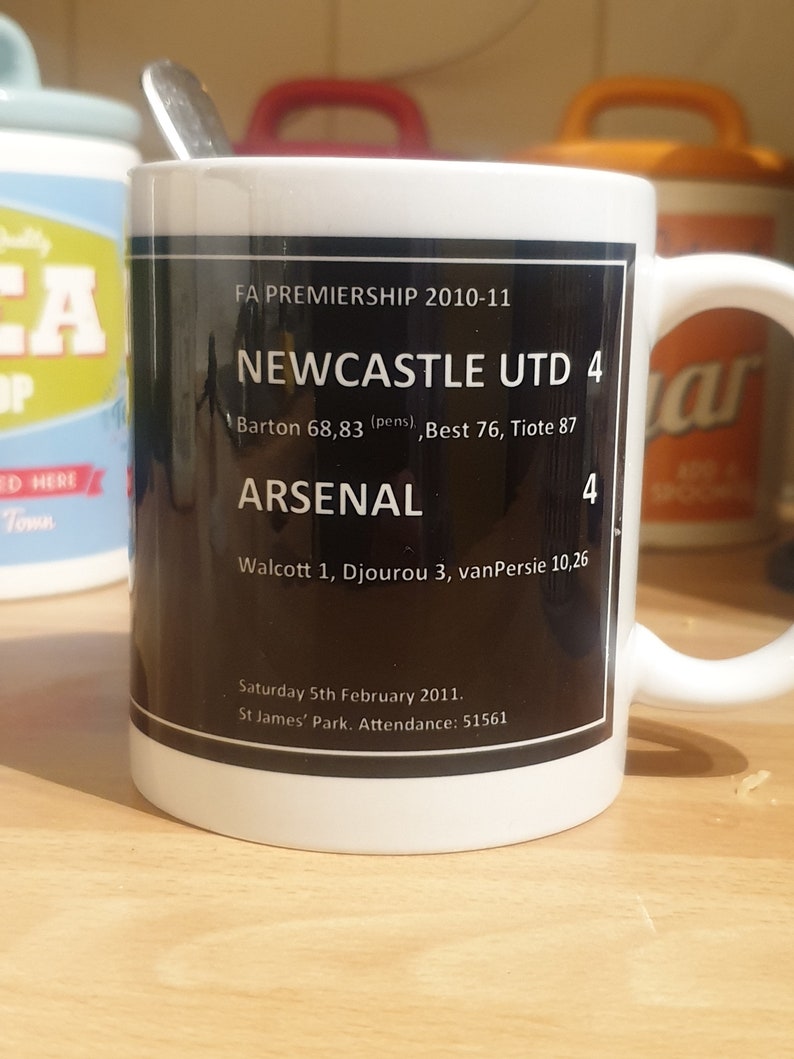 K&ouml;nnte beinhalten: Eine wei&szlig;e Keramiktasse mit einem schwarzen Design, das den Text "FA Premiership 2010-11", "Newcastle Utd 4", "Arsenal 4" und die Ergebnisse des Spiels zeigt. Der Text "Saturday 5th February 2011. St James' Park. Attendance: 51561" ist ebenfalls enthalten.