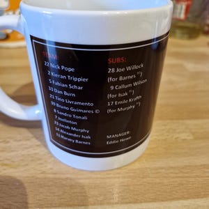 May include: A white ceramic mug with a black and red design. The mug features a list of soccer players and their numbers, including Nick Pope, Kieran Trippier, Fabian Schar, and Dan Burn. The mug also includes the team's manager, Eddie Howe.
