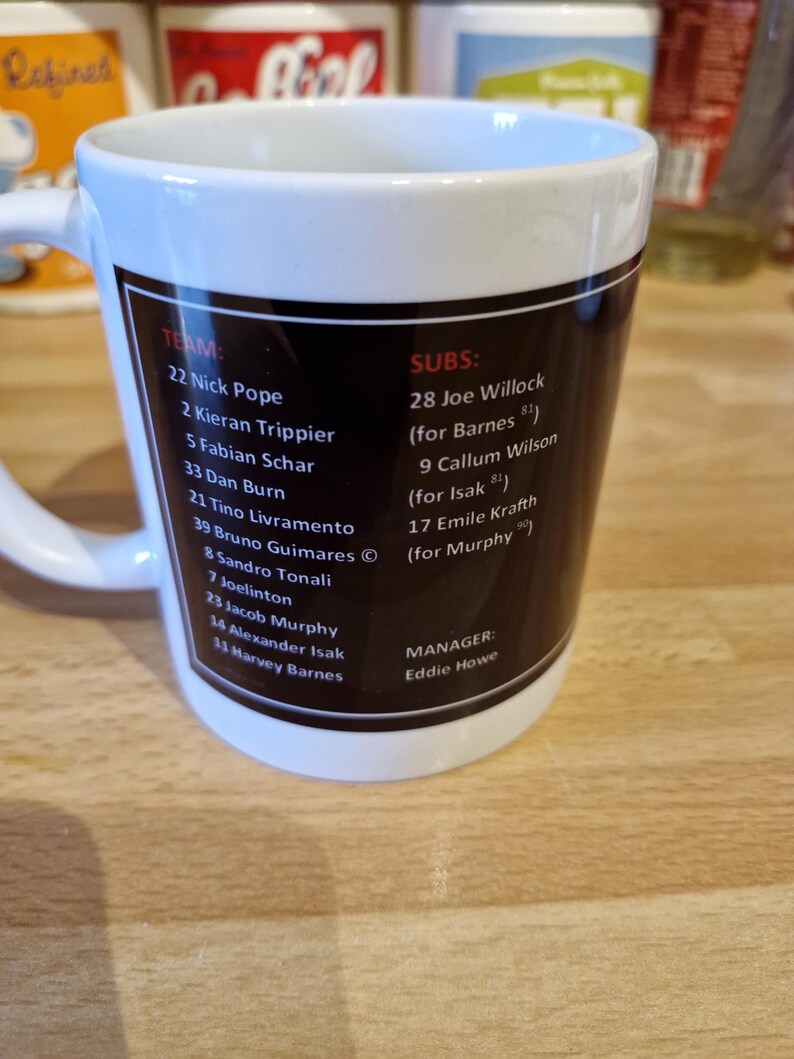 May include: A white ceramic mug with a black and red design. The mug features a list of soccer players and their numbers, including Nick Pope, Kieran Trippier, Fabian Schar, and Dan Burn. The mug also includes the team's manager, Eddie Howe.