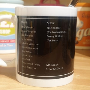 K&ouml;nnte beinhalten: Eine wei&szlig;e Keramiktasse mit einem schwarzen Band um die Mitte. Das Band hat wei&szlig;en Text, der "TEAM: Steve Harper, Danny Simpson, Jose Enrique, Kevin Nolan, Fabricio Coloccini, Mike Williamson, Joey Barton, Cheick Tiote, Leon Best, Peter Lovenkrands, Jonas Gutierrez" und "SUBS: Nile Ranger (for Lovenkrands), Danny Guthrie (for Best)" und "MANAGER: Steve McClaren" lautet.