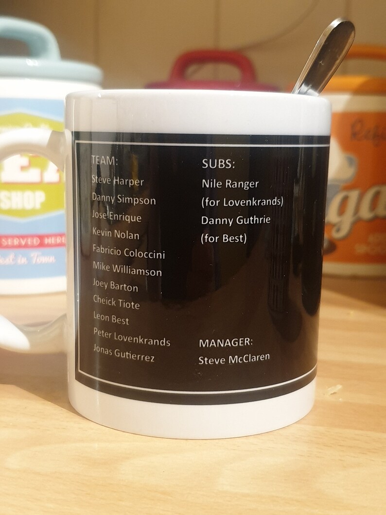 K&ouml;nnte beinhalten: Eine wei&szlig;e Keramiktasse mit einem schwarzen Band um die Mitte. Das Band hat wei&szlig;en Text, der "TEAM: Steve Harper, Danny Simpson, Jose Enrique, Kevin Nolan, Fabricio Coloccini, Mike Williamson, Joey Barton, Cheick Tiote, Leon Best, Peter Lovenkrands, Jonas Gutierrez" und "SUBS: Nile Ranger (for Lovenkrands), Danny Guthrie (for Best)" und "MANAGER: Steve McClaren" lautet.