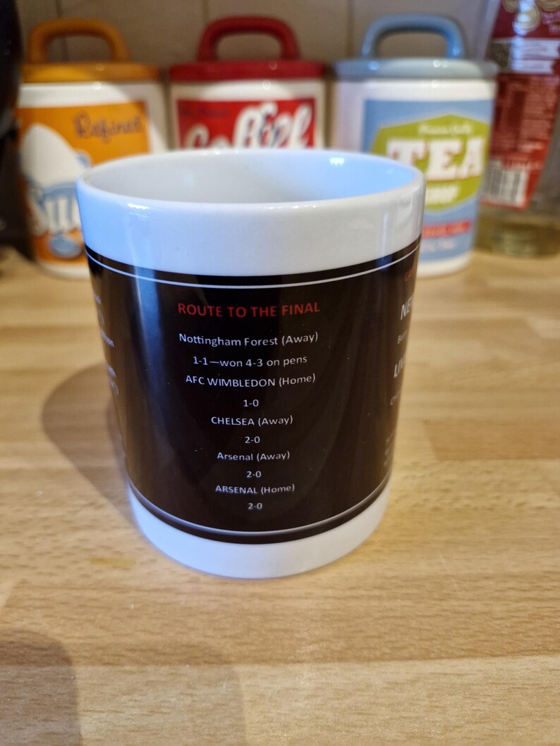 May include: A white ceramic mug with a black design that lists the route to the final of a soccer tournament. The text includes the teams and scores of each match. The mug is on a wooden surface.
