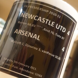 K&ouml;nnte beinhalten: Eine wei&szlig;e Keramiktasse mit einem schwarzen Design. Das Design zeigt den Text "FA Premiership 2010-11", "Newcastle Utd 4", "Arsenal 4", "Barton 68,83 (pens), Best 76, Tiote 87", "Walcott 1, Djourou 3, van Persie 10,26", "Samstag, 5. Februar 2011", "St James' Park. Zuschauer: 51561".