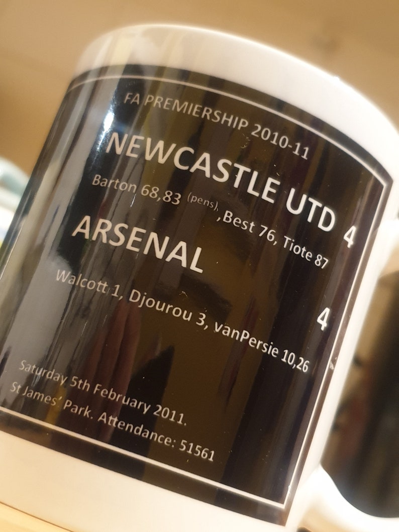 K&ouml;nnte beinhalten: Eine wei&szlig;e Keramiktasse mit einem schwarzen Design. Das Design zeigt den Text "FA Premiership 2010-11", "Newcastle Utd 4", "Arsenal 4", "Barton 68,83 (pens), Best 76, Tiote 87", "Walcott 1, Djourou 3, van Persie 10,26", "Samstag, 5. Februar 2011", "St James' Park. Zuschauer: 51561".