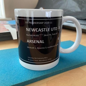 K&ouml;nnte beinhalten: Eine wei&szlig;e Keramiktasse mit einem schwarzen Design, das den Spielstand eines Fu&szlig;ballspiels zwischen Newcastle United und Arsenal zeigt. Der Spielstand ist Newcastle 4, Arsenal 4. Das Spiel fand am 5. Februar 2011 im St. James' Park mit einer Zuschauerzahl von 51.561 statt.