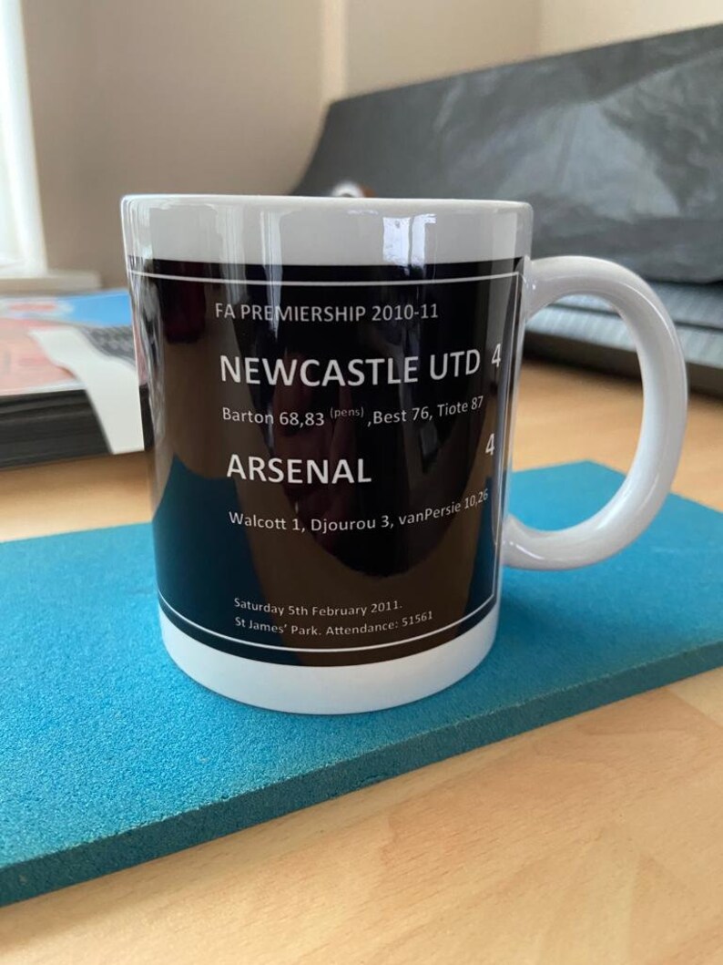 K&ouml;nnte beinhalten: Eine wei&szlig;e Keramiktasse mit einem schwarzen Design, das den Spielstand eines Fu&szlig;ballspiels zwischen Newcastle United und Arsenal zeigt. Der Spielstand ist Newcastle 4, Arsenal 4. Das Spiel fand am 5. Februar 2011 im St. James' Park mit einer Zuschauerzahl von 51.561 statt.