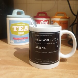 K&ouml;nnte beinhalten: Eine wei&szlig;e Keramiktasse mit einem schwarzen Design, das den Text "FA Premiership 2010-11", "Newcastle Utd 4", "Arsenal 4" sowie das Spielergebnis und das Datum zeigt. Die Tasse steht auf einer Holzoberfl&auml;che.