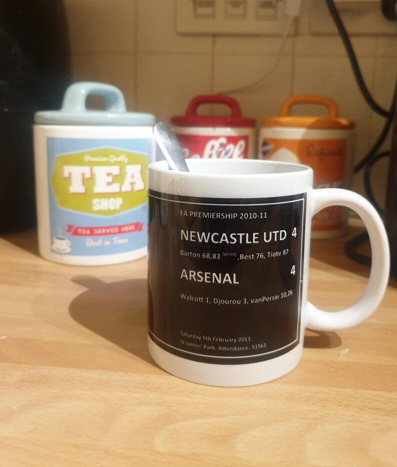 K&ouml;nnte beinhalten: Eine wei&szlig;e Keramiktasse mit einem schwarzen Design, das den Text "FA Premiership 2010-11", "Newcastle Utd 4", "Arsenal 4" sowie das Spielergebnis und das Datum zeigt. Die Tasse steht auf einer Holzoberfl&auml;che.