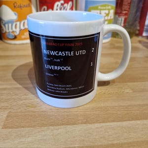 May include: A white ceramic mug with a black design commemorating the Carabao Cup Final 2025. The design includes the score of the game, Newcastle United 2, Liverpool 1, and the date and location of the game.