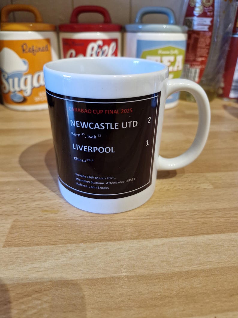 May include: A white ceramic mug with a black design commemorating the Carabao Cup Final 2025. The design includes the score of the game, Newcastle United 2, Liverpool 1, and the date and location of the game.