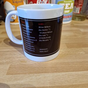 May include: A white ceramic mug with a black design featuring a soccer team roster. The mug has a black handle and a blue rim. The text on the mug includes the team name, player names, and numbers, as well as the manager's name, Eddie Howe.