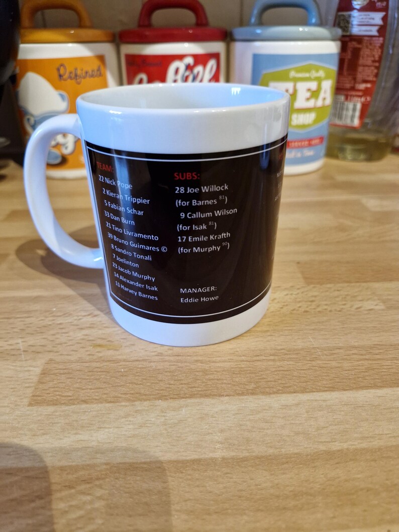 May include: A white ceramic mug with a black design featuring a soccer team roster. The mug has a black handle and a blue rim. The text on the mug includes the team name, player names, and numbers, as well as the manager's name, Eddie Howe.