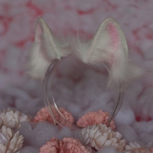 Puede incluir: Diadema de orejas de gato de pelo blanco con una diadema de plástico transparente. La diadema está sobre una superficie blanca y esponjosa con flores rosas.