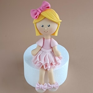 Pode incluir: Um topo de bolo de fondant rosa e branco com uma menina de cabelo loiro, um laço rosa e um vestido rosa. A menina está sentada em um suporte de bolo branco.
