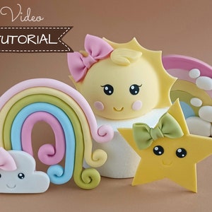 Peut inclure: Un ensemble de quatre décorations de gâteau en fondant représentant un soleil jaune avec un nœud rose, un arc-en-ciel avec un arc bleu, rose et jaune, un nuage blanc avec un nœud rose et une étoile jaune avec un nœud vert. Les décorations sont ornées d'yeux noirs et d'une bouche souriante.