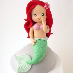 May include: A handcrafted mermaid figurine with red hair, a pink seashell top, and a green scaled tail. The mermaid has a small pink flower in her hair and is smiling. The figurine is on a gray base.