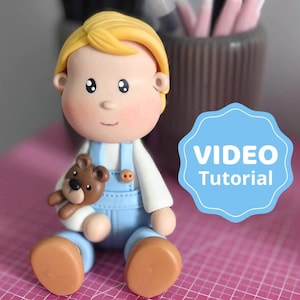 Puede incluir: Una figurita hecha a mano de un niño con cabello rubio, usando un overol azul y sosteniendo un oso de peluche marrón. La figurita está sentada sobre una superficie rosa con un letrero azul y blanco que dice "VIDEO Tutorial".