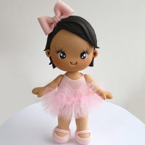 Puede incluir: Un adorno de pastel de bailarina rosa y marrón con un lazo rosa en el pelo. La bailarina lleva un tutú rosa y zapatos rosas.