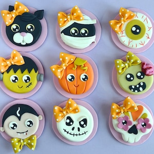 以下が含まれることがあります： フォンダンで飾られたハロウィンをテーマにしたケーキポップが9個。ケーキポップは、ミイラ、カボチャ、幽霊、吸血鬼、目など、さまざまなキャラクターを特徴としています。各ケーキポップは、オレンジ、イエロー、ホワイトのフォンダンでできたリボンで飾られています。