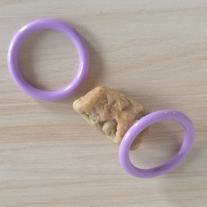 Op de afbeelding: Twee paarse plastic ringen op een licht houten oppervlak. Eén ring ligt plat, de andere staat rechtop met een kleine, lichtbruine koek erin.