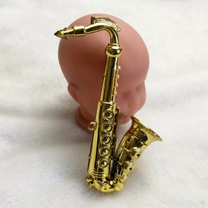 Puede incluir: Un saxofón de juguete de color dorado con un extremo en forma de campana. El saxofón está sobre una superficie blanca.