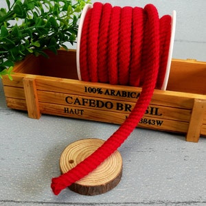 Puede incluir: Una bobina de cuerda de algodón roja, enrollada alrededor de un tubo de cartón blanco, se encuentra en una caja de madera con el texto "100% ARABICA CAFEDO BRASIL HAUT 3843W".