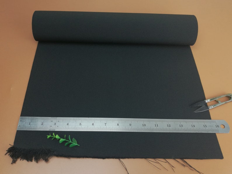 May include: Black fabric with a frayed edge. A ruler is placed on top of the fabric, with the numbers 1 through 16 visible. A small green plant is in the foreground.