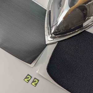 May include: Two pieces of black fabric with different textures. One piece has a smooth surface and the other has a textured surface. A silver iron is resting on top of the fabric.