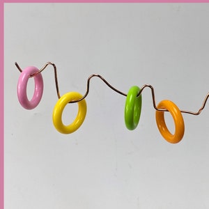 May include: Four colorful wooden rings hanging on a copper wire. The rings are pink, yellow, green, and orange.