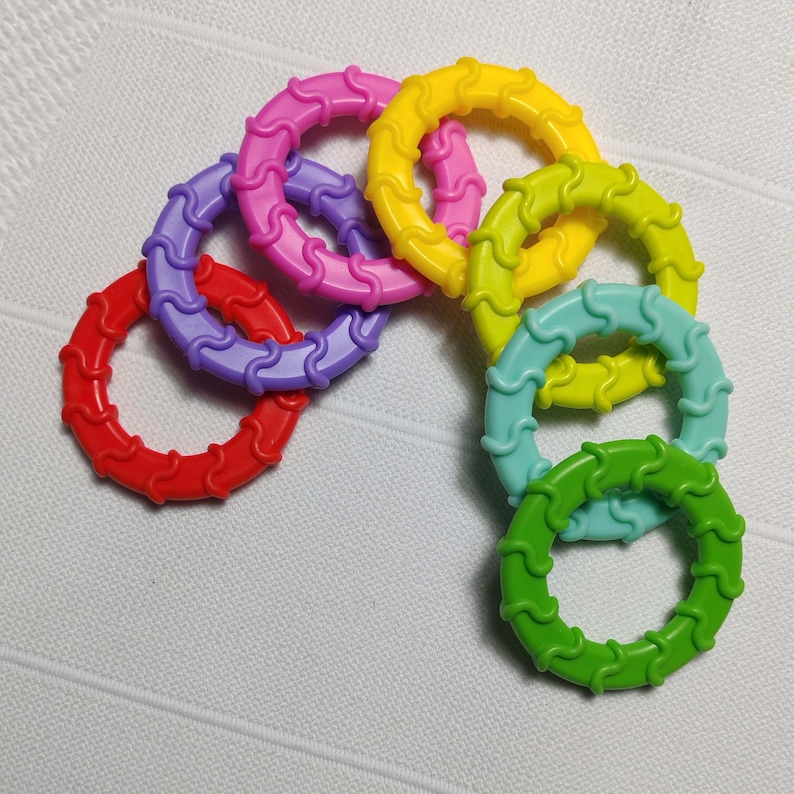 May include: A set of six colorful silicone teething rings in red, purple, yellow, green, blue, and pink. Each ring has a wavy texture and is designed for babies to chew on.
