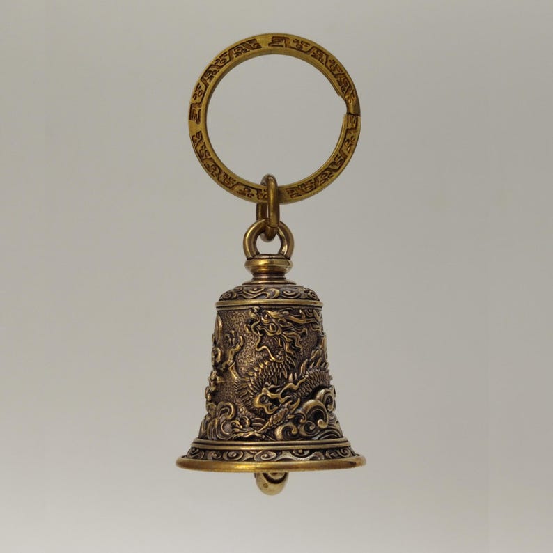 May include: A bronze-colored keychain featuring a bell-shaped charm. The bell is intricately detailed with a dragon design and ornamental patterns. The keychain includes a circular ring for attaching keys.