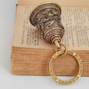 May include: A detailed brass bell keychain with an ornate dragon design. The bell is attached to a large, engraved ring. The bell is resting on an open book with Chinese characters.