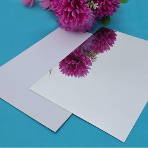 Könnte beinhalten: Zwei Blätter silbernes Spiegel-Acrylglas, übereinander gelegt, mit einem Strauß rosa Blumen, der sich im oberen Blatt spiegelt.