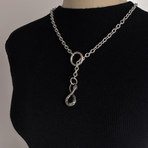 May include: Silver necklace with a chain, a large ring, and a snake pendant. The snake pendant is shaped like an infinity symbol. The necklace is worn against a black top.