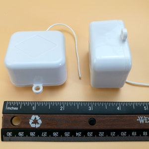 May include: Two white plastic music box mechanisms. One is square with a diamond design on top, the other is rectangular. A wooden ruler with inch measurements is in the foreground. The ruler shows measurements in cm.