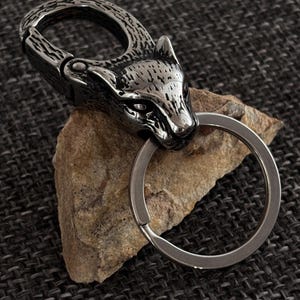 Può includere: Un portachiavi color argento con un design a testa di lupo. Il portachiavi ha un moschettone a molla e un anello portachiavi. La testa di lupo è dettagliata con pelliccia e tratti del viso strutturati. Il portachiavi è in metallo.