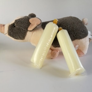 Op de afbeelding: Twee lange, cilindrische, lichtgele objecten in een doorzichtige plastic verpakking liggen voor een pluchen varkensspeelgoed. Het varken is grijs en wit met roze oren en snuit. De objecten lijken kaarsen of iets dergelijks te zijn.