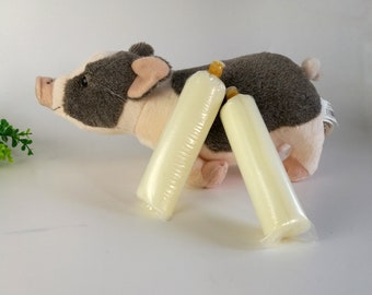 May include: Two long, cylindrical, pale yellow objects in clear plastic packaging are in front of a plush pig toy. The pig is gray and white with pink ears and snout. The objects appear to be candles or similar items.