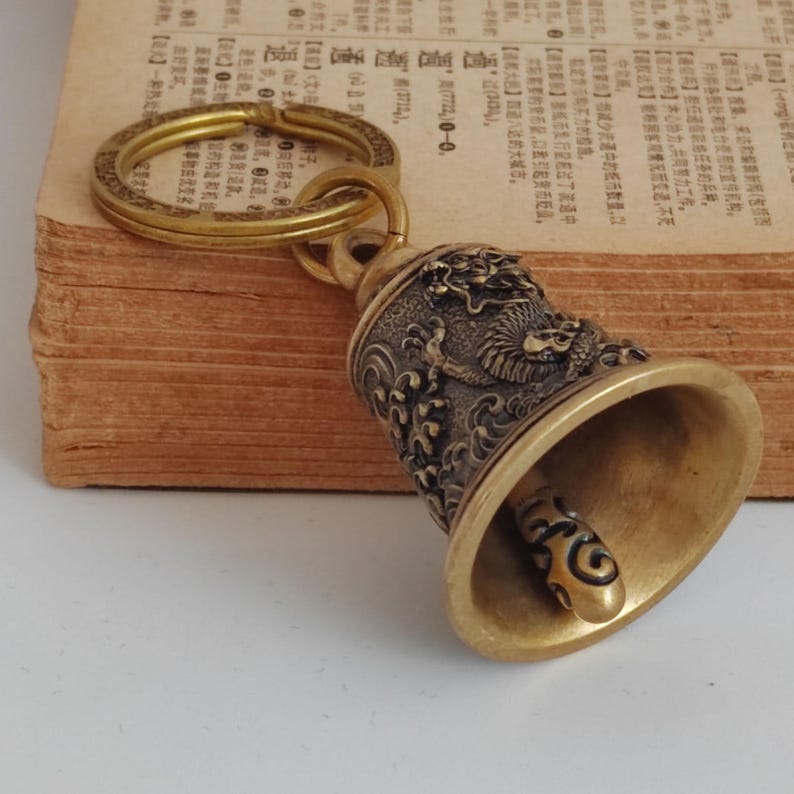 May include: A brass bell keychain with a dragon design. The bell has intricate details and a golden hue. The keychain is attached to a gold-colored ring, and the bell is approximately 2 inches tall.